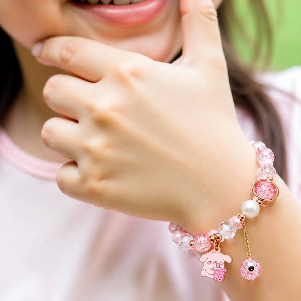 A close-up of a child's wrist wearing a pink beaded charm bracelet with a rabbit charm and a round pink bead, shown against a soft pink shirt and blurred green background.
